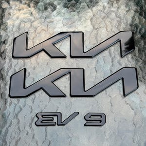 Könnte beinhalten: Zwei verchromte Kia-Embleme und ein passendes EV9-Emblem. Die Kia-Embleme sind das stilisierte Logo, und das EV9-Emblem ist eine Modellbezeichnung. Alle Embleme haben eine glänzende Oberfläche auf strukturiertem Hintergrund.