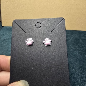 May include: A pair of small, pink paw print stud earrings. The earrings are set on a black earring display card. The paw prints are light pink with a clear border. The background is a neutral color.