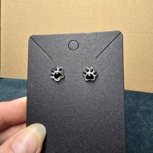 May include: A pair of silver-colored stud earrings shaped like paw prints. Each earring has a black enamel fill in the paw pads. The earrings are displayed on a black earring card.