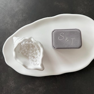 Könnte beinhalten: Eine weiße, muschelförmige Keramikschale mit einer weißen Perlenkette, begleitet von einer rechteckigen grauen Samt-Ringbox mit den in weißem Faden gestickten Buchstaben S & T auf dem Deckel, alles auf einem glatten, weißen, ovalen Tablett.