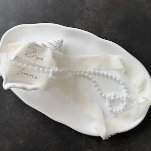 Könnte beinhalten: Eine ovale weiße Keramikschale mit einer weißen Muschel, einem cremefarbenen Band mit ausgefransten Kanten und den aufgedruckten Namen Besjan und Leonora sowie einer langen Kette aus weißen Kunstperlen, die locker auf der Schale liegt.