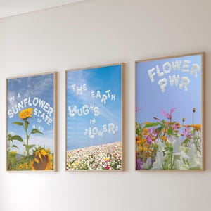 Könnte beinhalten: Drei gerahmte Kunstdrucke mit floralen Themen. Der linke Druck zeigt eine Sonnenblume mit dem Text "IN A SUNFLOWER STATE OF MIND". Der mittlere Druck zeigt ein Blumenfeld mit dem Text "THE EARTH LAUGHS IN FLOWERS". Der rechte Druck zeigt Blumen mit dem Text "FLOWER PWR".