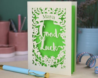 May include: A cream-colored laser-cut greeting card featuring a floral border and the text "Maya Good Luck" in a script font. The card has a bright green inner layer visible through the cutouts and stands upright on a flat surface.