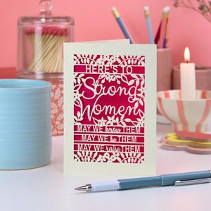 K&ouml;nnte beinhalten: Eine cremefarbene Gru&szlig;karte mit einem lasergeschnittenen Blumenrand und dem Text: "HERE'S TO Strong Women MAY WE know THEM MAY WE be THEM MAY WE raise THEM." Der Ausschnitt gibt einen leuchtend magentafarbenen Hintergrund frei.
