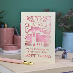 A laser cut Christening card with a church surrounded by trees, flowers, clouds and the sun. With a banner flying above the church for the Christening date and beneath the text "On Your Christening Day" and the child's name. Shown standing on a white desk with stationery items, laser cut from cream cardstock with a candy pink insert paper