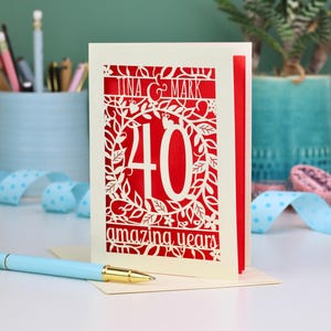 May include: A cream-colored greeting card with intricate laser-cut details featuring a red background. The card displays the names Tina & Mark, the number 40, and the text amazing years surrounded by a decorative floral and vine pattern.