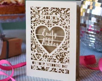 May include: A white laser-cut wedding card featuring an intricate floral and heart pattern. The text reads 24.08.2026, Emily and Ashley, and ON YOUR WEDDING DAY. The card has a tan inner layer visible through the cutouts and stands upright.