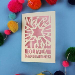 May include: A cream-coloured laser-cut greeting card featuring a Star of David, sunburst rays, and stars. The text reads "Mazel Tov!", "OLIVIA", and "ON YOUR BAT MITZVAH". The card is set against a blue background with colourful yarn pom-poms.