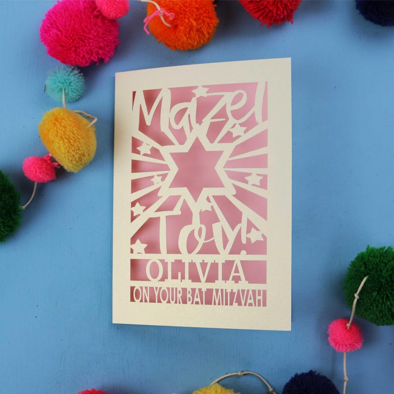 May include: A cream-coloured laser-cut greeting card featuring a Star of David, sunburst rays, and stars. The text reads "Mazel Tov!", "OLIVIA", and "ON YOUR BAT MITZVAH". The card is set against a blue background with colourful yarn pom-poms.