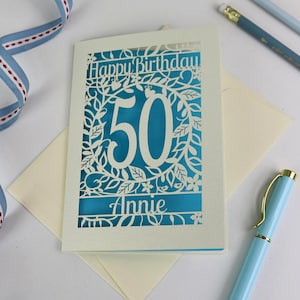 A laser cut flower birthday card with cut out floral design and "Happy Birthday" text. The birthday card is personalised with a cut out name and the age in the centre, surrounded by leaves and flowers