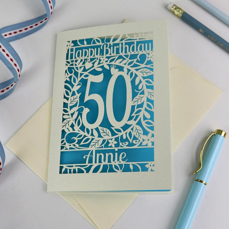 A laser cut flower birthday card with cut out floral design and "Happy Birthday" text. The birthday card is personalised with a cut out name and the age in the centre, surrounded by leaves and flowers