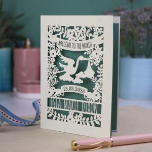 A gorgeous new baby card depicting a stork carrying a bundle surrounded with leaves, flowers and clouds. A banner saying Welcome To The World across the top,  and a second banner below with the baby's date of birth and their name under that. Shown standing on a white desk with stationery items in the fore and background. Its cut from cream cardstock and has a sage insert paper.