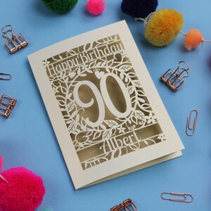 A laser cut keepsake 90th birthday card, personalised with a name in the bottom line of text. Card says Happy Birthday, 90. There is a floral border. Card is cream with a gold leaf insert