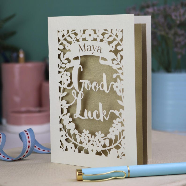 This floral Good Luck card is shown standing on a book with a pen, ribbon and ceramics in the background. It shows the text Good Luck in the centre of the card with a banner above showing the name Maya. It is laser cut from cream card with a gold leaf insert paper