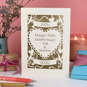 A laser cut wedding anniversary card with cut out heart design and "1976! in a banner, "Happy 50th Anniversary Val & Martin" in a heart and "2026" in a banner underneath