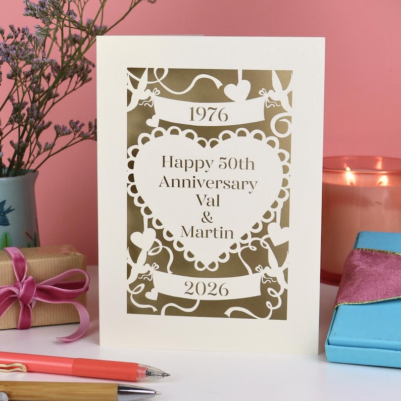 A laser cut wedding anniversary card with cut out heart design and "1976! in a banner, "Happy 50th Anniversary Val & Martin" in a heart and "2026" in a banner underneath