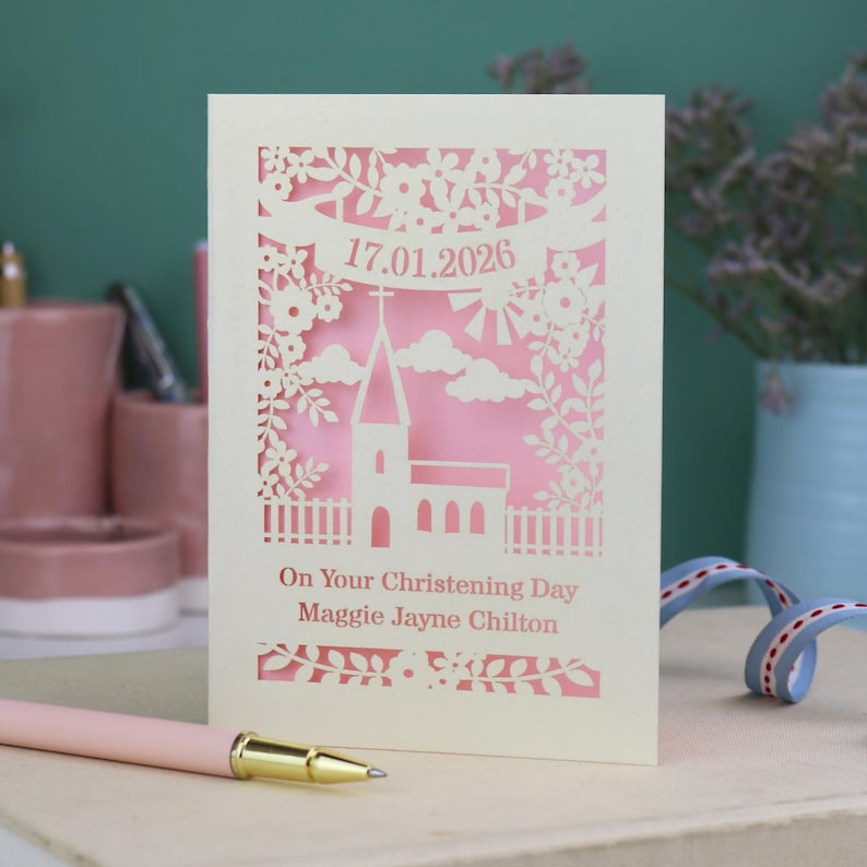 This beautiful laser cut Christening card shows a church surrounded by trees, flowers, clouds and the sun. With a banner flying above the church for the Christening date and beneath the text "On Your Christening Day" and the child's name. Shown standing on a white desk with stationery items, laser cut from cream cardstock with a candy pink insert paper
