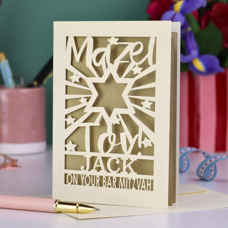 K&ouml;nnte beinhalten: Eine cremefarbene, lasergeschnittene Gru&szlig;karte mit einem Davidstern, Sternen und dem Text Mazel Tov! JACK ON YOUR BAR MITZVAH. Die Karte hat eine goldfarbene Innenschicht, die durch die filigranen Ausschnitte sichtbar ist.