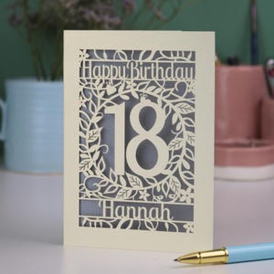 A laser cut flower birthday card with cut out floral design and "Happy Birthday" text. The birthday card is personalised with a cut out name and the age in the centre, surrounded by leaves and flowers