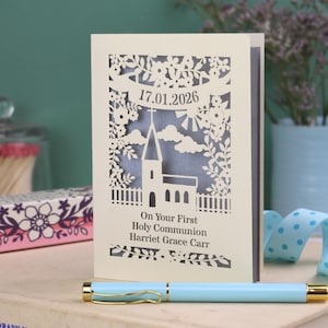 This beautiful laser cut First Holy Communion card shows a church surrounded by trees, flowers, clouds and the sun. With a banner flying above the church for the  Communion date and beneath the text "On Your First Holy Communion" and the child's name. Shown standing on a white desk with stationery items, laser cut from cream cardstock with a silver insert paper