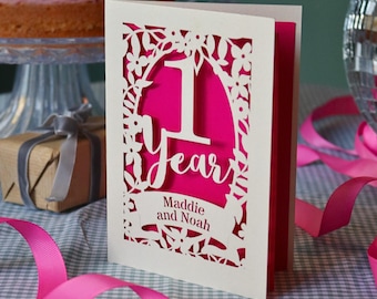 May include: A cream-colored greeting card with a laser-cut floral border and the text 1 Year, featuring the names Maddie and Noah printed below. The card has a vibrant hot pink inner layer visible through the intricate cutouts.