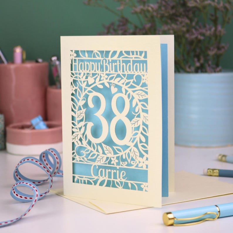 A paper cut flower birthday card with cut out floral design and "Happy Birthday" text. The birthday card is personalised with a cut out name and the age in the centre, surrounded by leaves and flowers