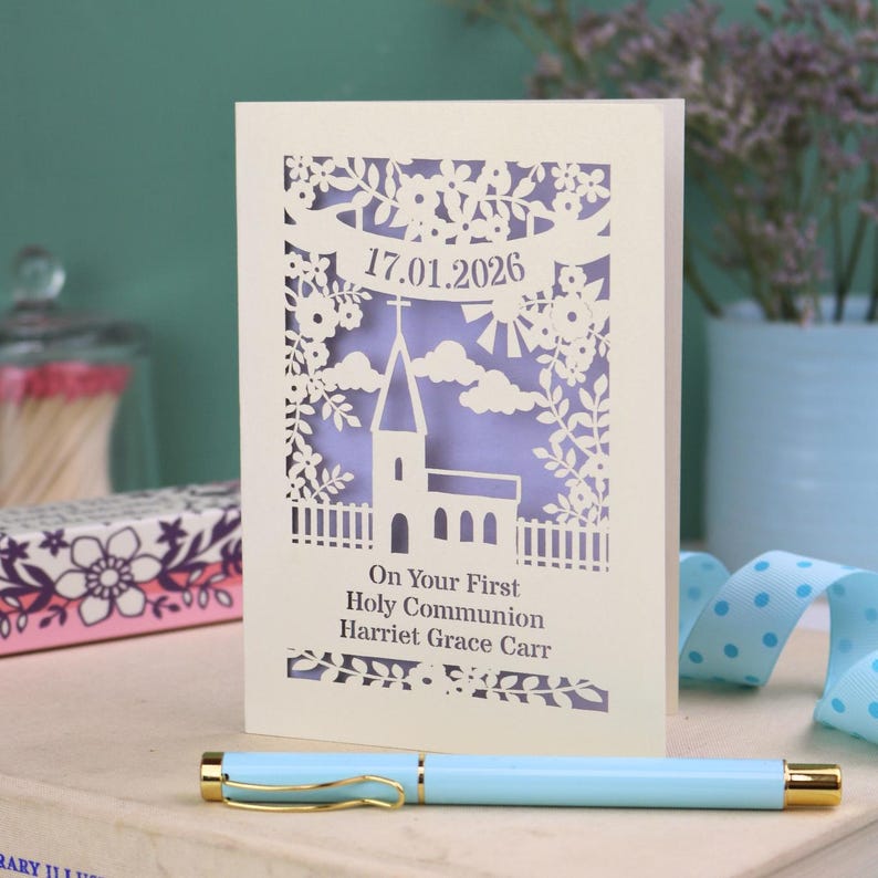 This beautiful laser cut First Holy Communion card shows a church surrounded by trees, flowers, clouds and the sun. With a banner flying above the church for the  Communion date and beneath the text "On Your First Holy Communion" and the child's name. Shown standing on a white desk with stationery items, laser cut from cream cardstock with a lilac insert paper