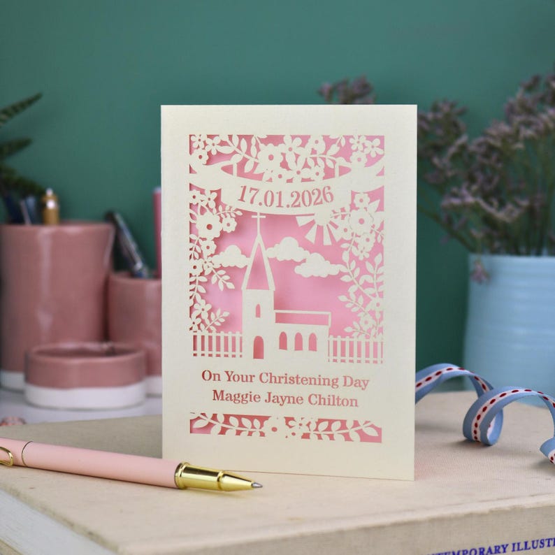 A laser cut Christening card with a church surrounded by trees, flowers, clouds and the sun. With a banner flying above the church for the Christening date and beneath the text "On Your Christening Day" and the child's name. Shown standing on a white desk with stationery items, laser cut from cream cardstock with a candy pink insert paper