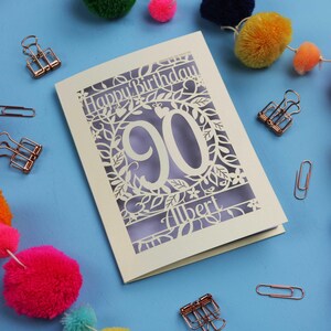 A laser cut keepsake 90th birthday card, personalised with a name in the bottom line of text. Card says Happy Birthday, 90. There is a floral border. Card is cream with a lilac insert
