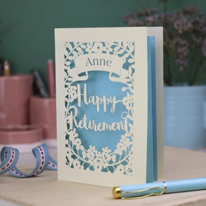 This lovely laser cut retirement card is shown standing on a white desk with a pen and ceramics in the background. It has the words Happy Retirement cut in a script font, has a banner above for the retiree's name and a floral decorative border. It is cut from cream cardstock and has a light blue insert paper