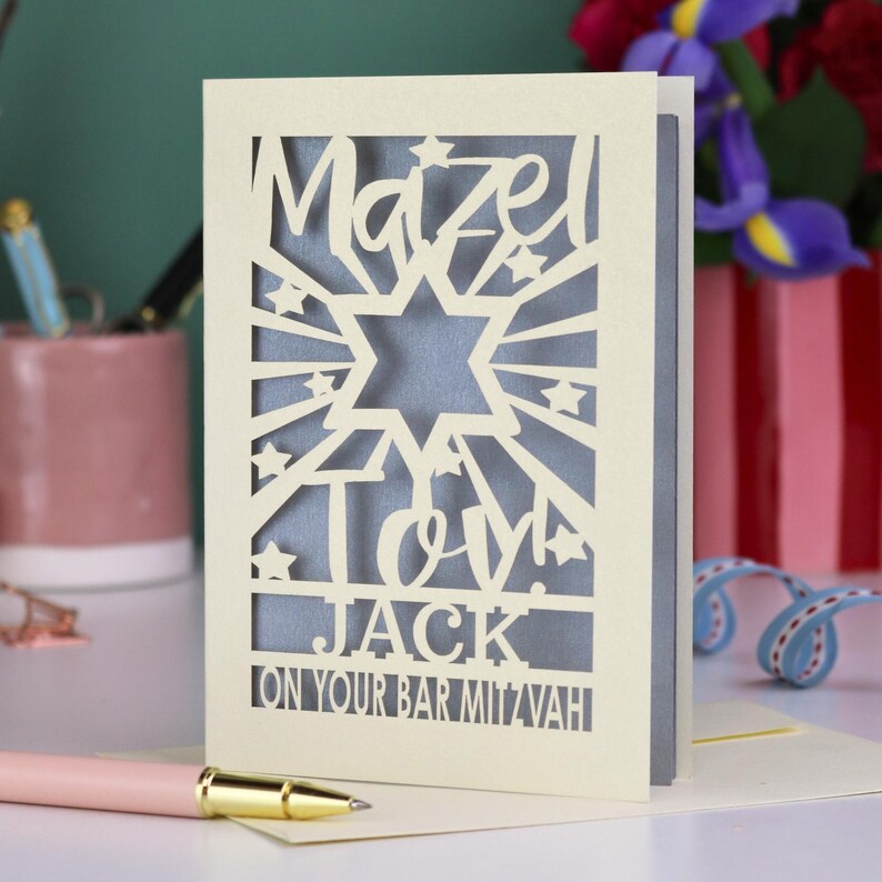 K&ouml;nnte beinhalten: Eine cremefarbene, lasergeschnittene Gru&szlig;karte mit einem Davidstern, Sternakzenten und dem Text "Mazel Tov! JACK ON YOUR BAR MITZVAH" in einer kr&auml;ftigen Stanzschrift. Durch die filigranen Ausschnitte ist ein grauer Innenteil zu sehen.