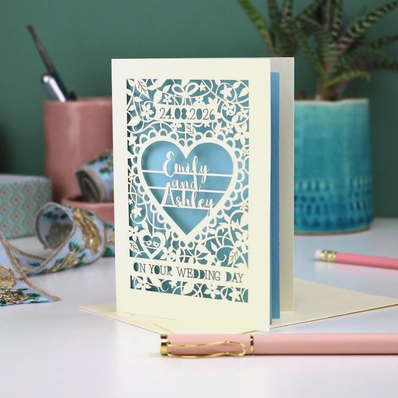 A laser cut "on your wedding day" card with floral design and cut out personalised text. Card features the wedding date, the first names of the couple in a heart shape, and the text "On your wedding day" Standing on a white desk with stationery items in the background. Cut from cream card stock with a light blue insert paper