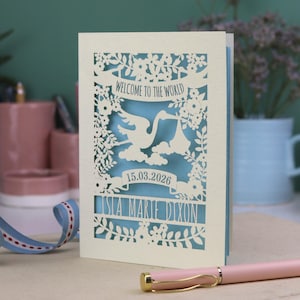 A gorgeous new baby card depicting a stork carrying a bundle surrounded with leaves, flowers and clouds. A banner saying Welcome To The World across the top,  and a second banner below with the baby's date of birth and their name under that. Shown standing on a white desk with stationery items in the fore and background. Its cut from cream cardstock and has a light blue insert paper.
