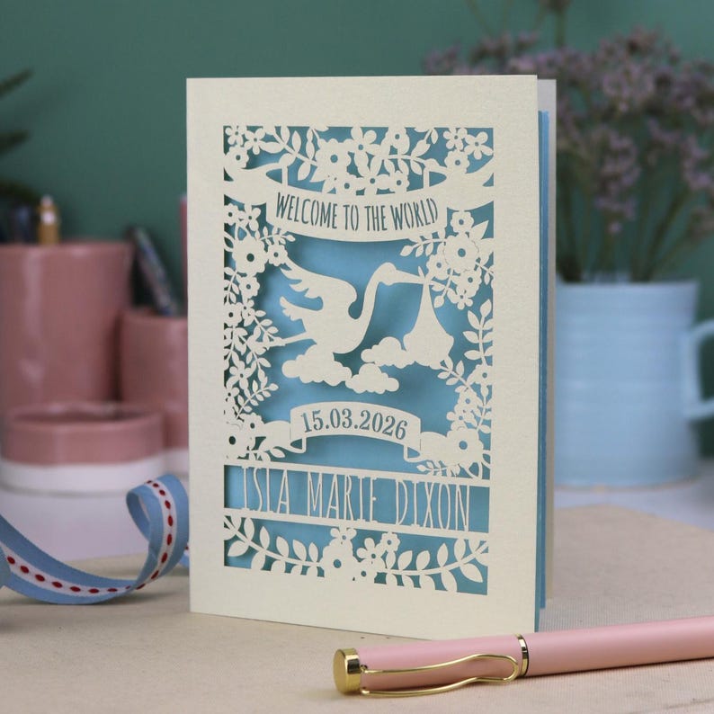 A gorgeous new baby card depicting a stork carrying a bundle surrounded with leaves, flowers and clouds. A banner saying Welcome To The World across the top,  and a second banner below with the baby's date of birth and their name under that. Shown standing on a white desk with stationery items in the fore and background. Its cut from cream cardstock and has a light blue insert paper.