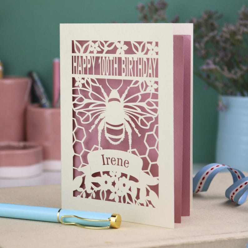 Peut inclure: Une carte de v&oelig;ux d&eacute;coup&eacute;e au laser de couleur cr&egrave;me avec un motif d'abeille et de nid d'abeille, portant le texte "HAPPY 100TH BIRTHDAY" et le nom "Irene" sur une banni&egrave;re. Un insert rose poudr&eacute; est visible &agrave; travers les d&eacute;coupes.