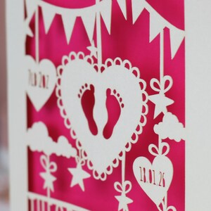 A close up view of the details on a laser cut new baby card  This cute card is laser cut from a cream cardstock with a shocking pink paper insert and features a heart with a pair of baby feet cut out, bunting, clouds, stars and hearts. The text under the heart is the baby's name Theodore Lucas and the left hanging heart shows the baby's weight, 7lb 2oz, and the right hanging heart shows the baby's date of birth 18/01/26