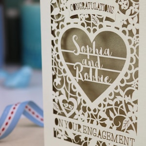 A laser cut engagement card with intricate floral cut out design, the word "Congratulations" in a banner at the top, a heart shape border with 3 lines of text, 'Name and Name', and 'On Your Engagement' in cut out lettering across the bottom of the card.