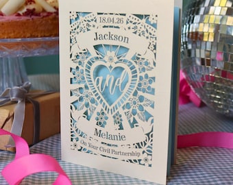 Puede incluir: Una tarjeta de felicitación de color crema cortada con láser que presenta un corazón central, motivos florales y el texto 18.04.26, Jackson and Melanie, On Your Civil Partnership, sobre un fondo azul claro con bordes decorativos.