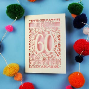 May include: A cream-colored card with a laser-cut design, featuring the words "MUM & DAD" and "60 amazing years." The number 60 is prominently displayed within a floral wreath. Colorful pom-pom garland decorates the blue background.