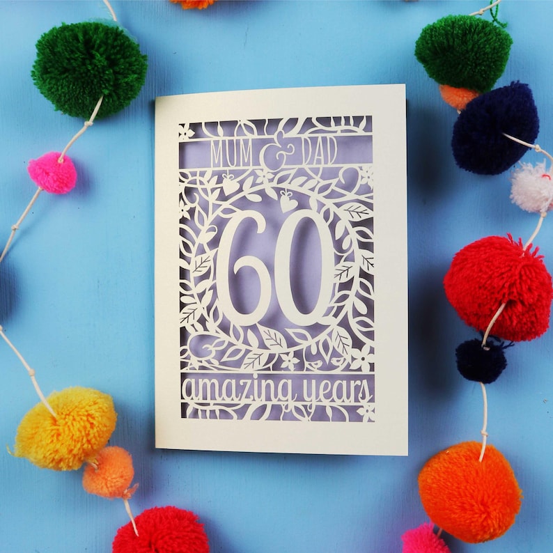 May include: A cream-colored 60th anniversary card with intricate cut-out floral and leaf designs. The card reads "MUM & DAD" at the top and "amazing years" at the bottom. Colorful pom-pom garland decorates the background.