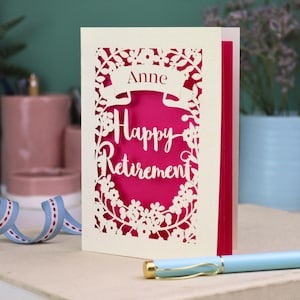 This lovely laser cut retirement card is shown standing on a white desk with a pen and ceramics in the background. It has the words Happy Retirement cut in a script font, has a banner above for the retiree's name and a floral decorative border. It is cut from cream cardstock and has a shocking pink insert paper