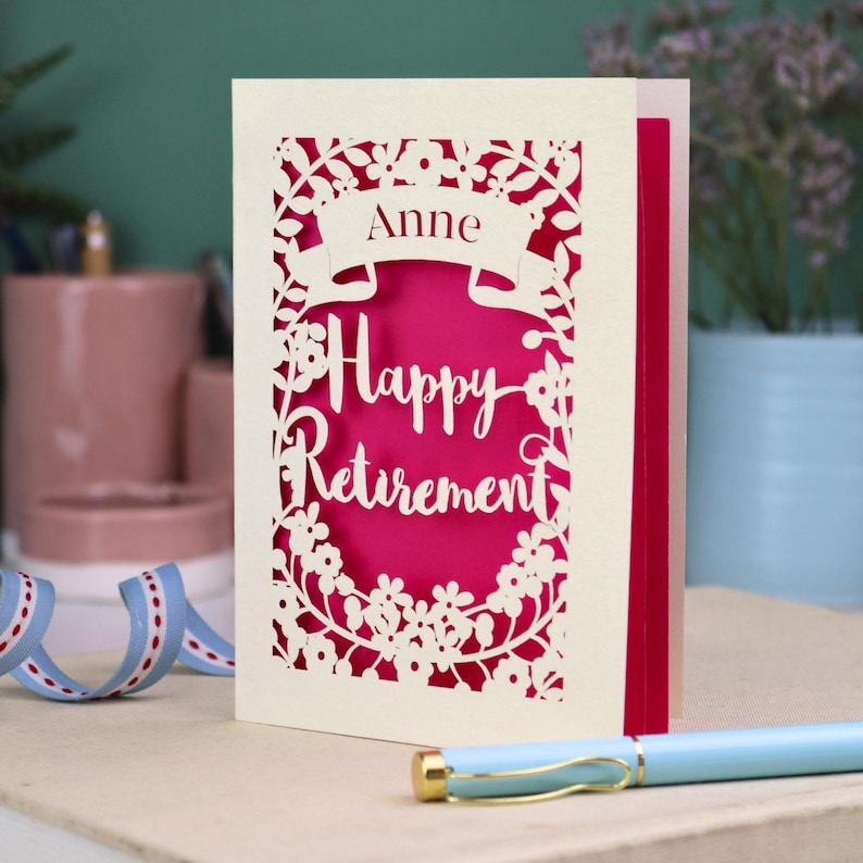 This lovely laser cut retirement card is shown standing on a white desk with a pen and ceramics in the background. It has the words Happy Retirement cut in a script font, has a banner above for the retiree's name and a floral decorative border. It is cut from cream cardstock and has a shocking pink insert paper