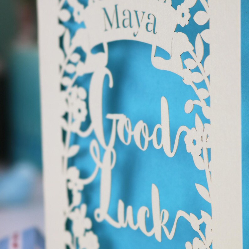 A close up detail of the text on this good luck card showing the text Good Luck and the name Maya cut from cream card with a peacock blue insert paper
