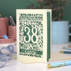A laser cut flower birthday card with cut out floral design and "Happy Birthday" text. The birthday card is personalised with a cut out name and the age in the centre, surrounded by leaves and flowers
