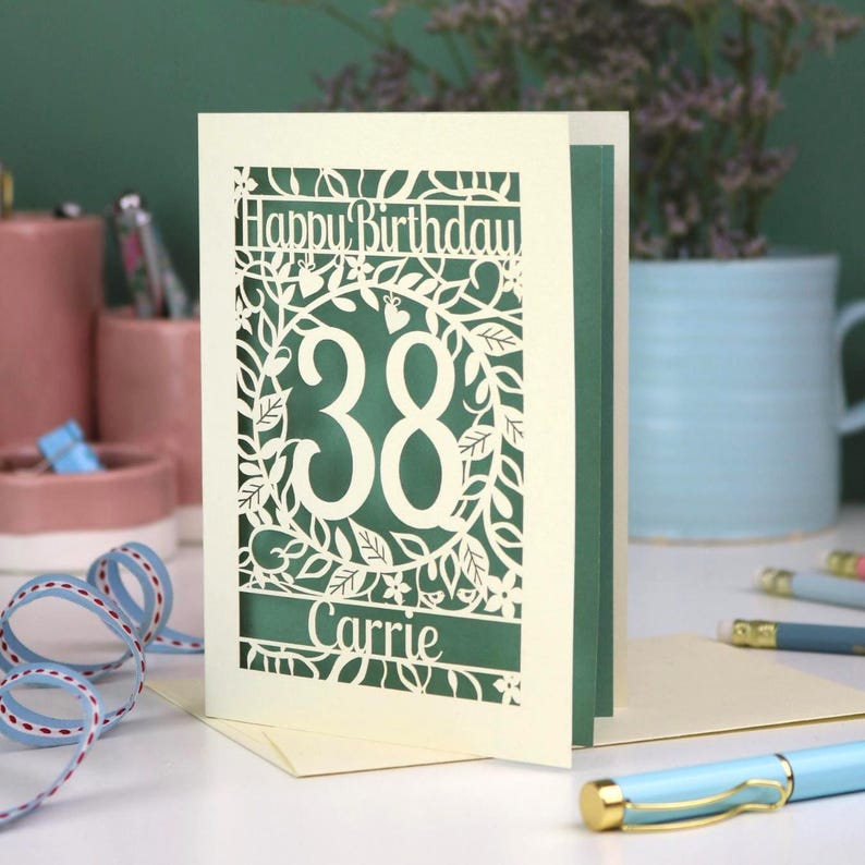 A laser cut flower birthday card with cut out floral design and "Happy Birthday" text. The birthday card is personalised with a cut out name and the age in the centre, surrounded by leaves and flowers