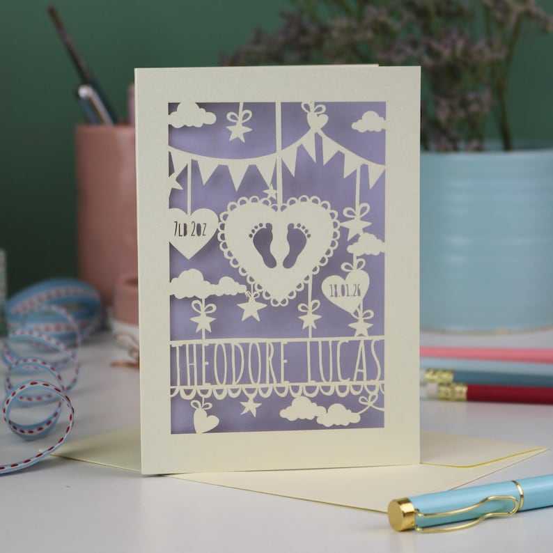 A laser cut new baby card on a white desk with stationery items in the 
background. This cute card is laser cut from a cream cardstock with a lilac paper insert and features a heart with a pair of baby feet cut out, bunting, clouds, stars and hearts. The text under the heart is the baby's name Theodore Lucas and the left hanging heart shows the baby's weight, 7lb 2oz, and the right hanging heart shows the baby's date of birth 18/01/26