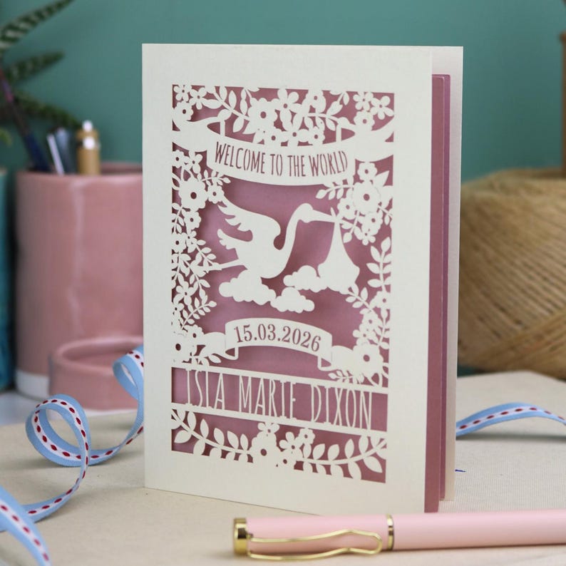A gorgeous new baby card depicting a stork carrying a bundle surrounded with leaves, flowers and clouds. A banner saying Welcome To The World across the top,  and a second banner below with the baby's date of birth and their name under that. Shown standing on a white desk with stationery items in the fore and background. Its cut from cream cardstock and has a dusky pink insert paper.