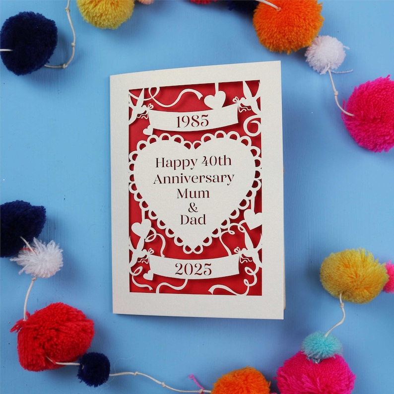 A personalised 40th wedding anniversary card, laser cut from a cream card with red paper backing. The year of wedding and year of anniversary feature in the top and bottom banners. There is a heart shape in the centre of the design with text cut out