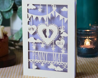 May include: A cream-colored laser-cut greeting card featuring a central heart with baby footprints, hanging stars, clouds, and bunting. The text reads "6lb 9oz", "18.02.26", and "PENNY MAE" against a light purple background.