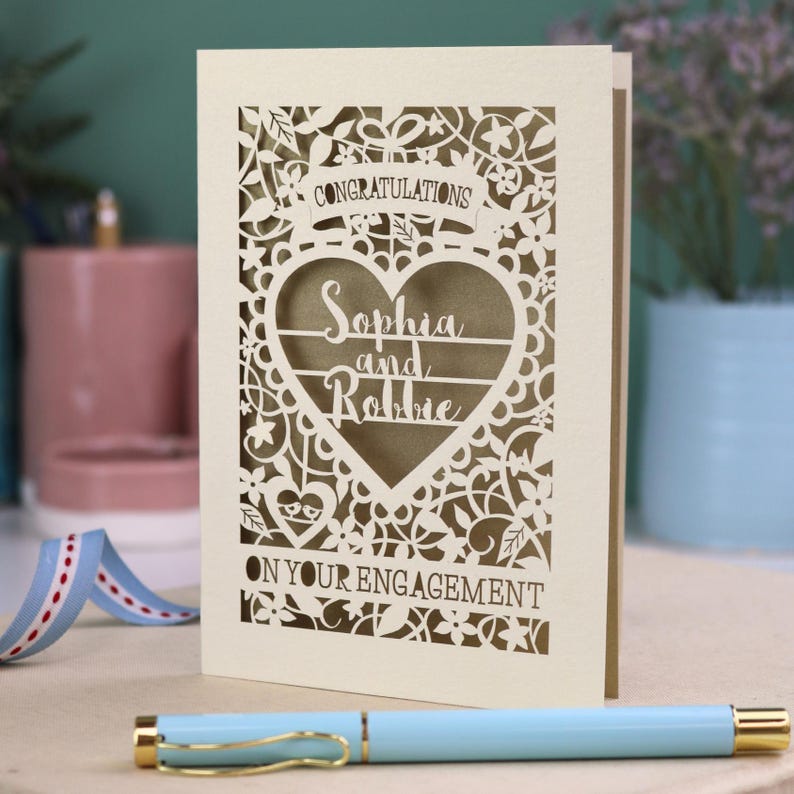 A laser cut engagement card with intricate floral cut out design, the word "Congratulations" in a banner at the top, a heart shape border with 3 lines of text, 'Name and Name', and 'On Your Engagement' in cut out lettering across the bottom of the card.